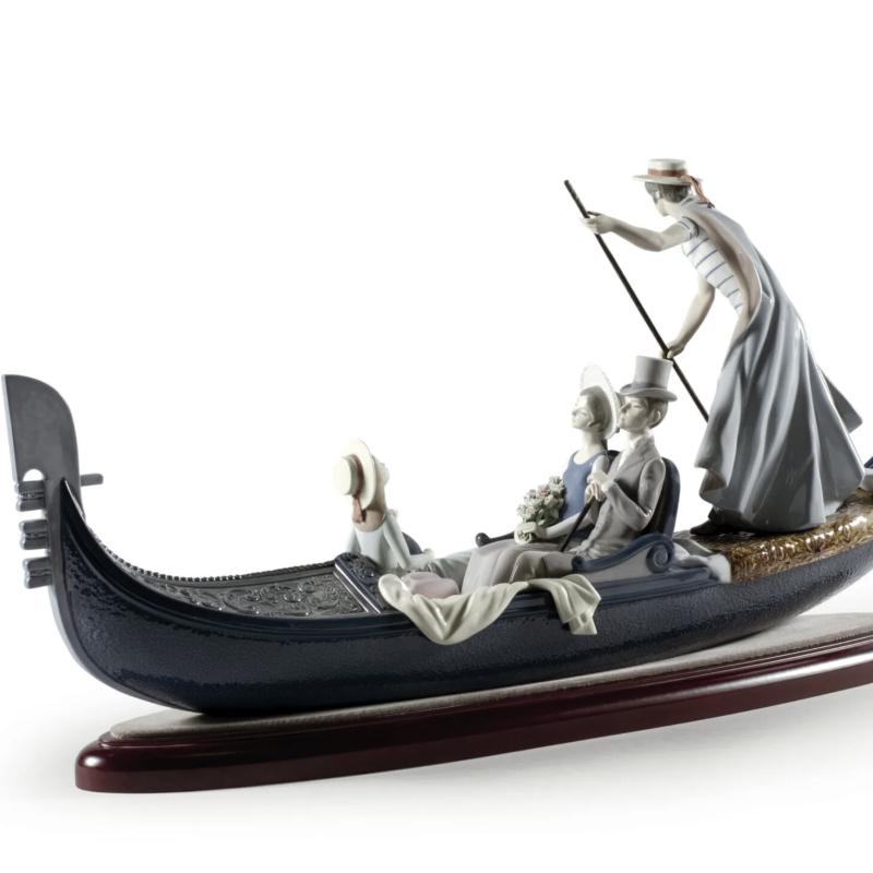 In The Gondola Couple Sculpture. Numbered Edition  Numbered glossy porcelain sculpture of a gondola with young bride and groom enjoying a walk and gondolier in typical striped t-shirt and straw hat attire. 01001350